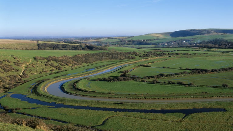 A wide, sweeping view of a green river valley with a winding river cutting through open fields. Low, gently rolling hills stretch into the distance under a clear blue sky, with patches of shrubs and hedgerows scattered across the landscape